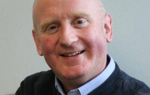 Peter is a white man with a bald head and wearing a pale blue shirt under a navy jumper. He is smiling.