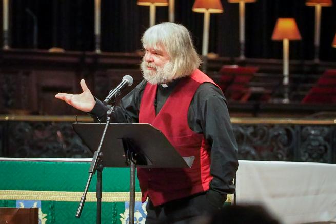 Malcolm Guite stands on the dais at St Paul's Cathedral, hand raised as he speaks, green altar cloth on the altar behind him.