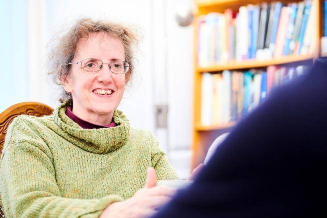 Karen is a white woman with light wavy hair pinned up and glasses, wearing a pale green rollneck sweater, smiling at someone to the side of the camera.