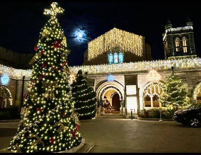 The Cathedral Church lit up with Christmas lights, with a Christmas tree in the foreground also decorated with lights and a cross on top. The sky is dark and a full moon can be seen above the Church.