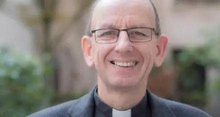 Neil is a white man with balding grey hair and wire framed glasses, wearing a black clerical shirt and black jacket. He smiles at the camera, trees and a garden wall blurred in the background.