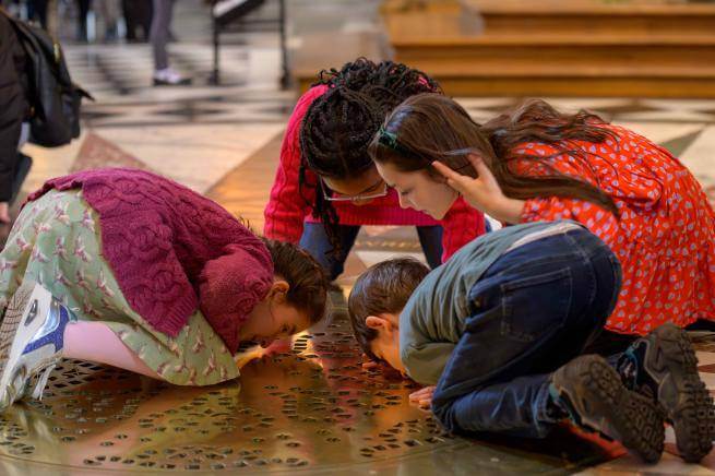 Four young children kneel on the floor of St Paul's Cathedral, with their faces close to the ground, looking through a golden grille through to the Cathedral Crypt.