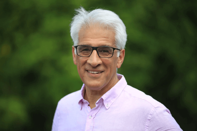 Steve is a mixed race man with white hair and black glasses, wearing a pink shirt, and standing outside with out of focus green foliage behind him.