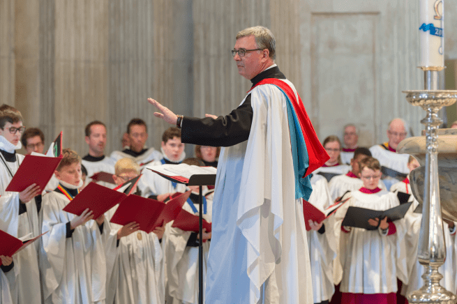 A white man with grey hair and glasses wearing a choir robe conducts a choir of choristers, also wearing white robes.