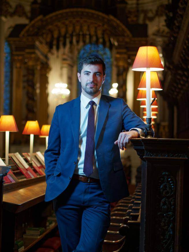 A portrait of James Orford. James is a white man with dark, short hair and a beard. He is wearing a suit and tie, and is stood in the Quire of St Paul's Cathedral.