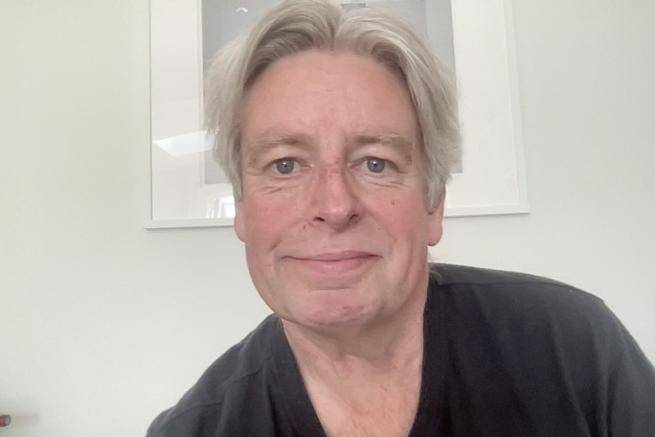 Graeme is a white man with short grey hair and a black top, seated in front of a white wall with a white framed photo behind him.