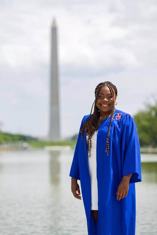 Alexandra is a young black woman wearing an academic robe standing in front of the Washington Monument and Lincoln Memorial Pool