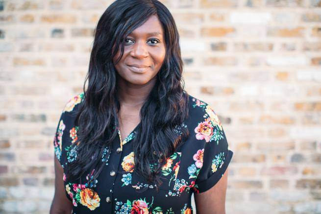 Chine is a black woman with long black hair wearing a floral patterned black dress and standing in front of a brick wall