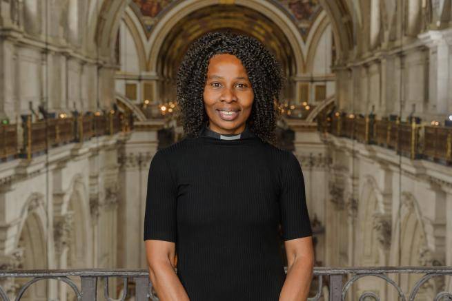 Catherine Okoronkwo is a black woman with shoulder length braided hair, wearing a short sleeved black top with a clerical collar. She stands in the Cathedral high up on the triforium level, with the nave of the Cathedral behind her.