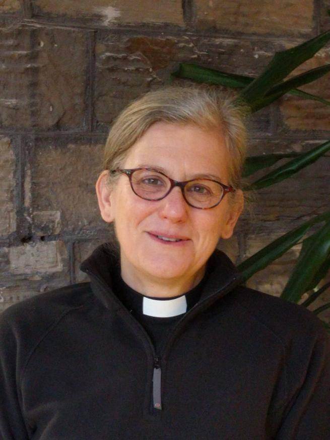 Janet Williams is a white woman with short grey hair and dark rimmed glasses wearing a clerical collar under a black jacket