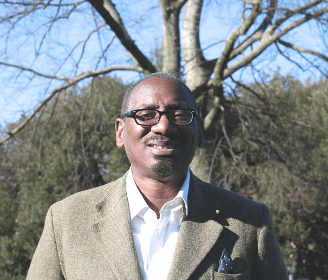 Richard is a black man with a bald head and dark rimmed glasses wearing a beige suit jacket over a white shirt, standing outdoors in front of a bare tree and a pale blue sky