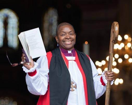 Bishop Rose is a black woman with sort hair wearing a bishop's robes holding an order of service up in one hand and a bishop's crook in the other, she is emerging from a building with candle light in the background, and smiling to the camera