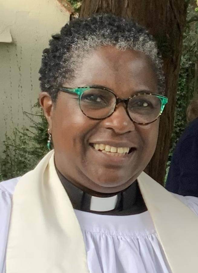 Ade is a black woman with dark hair greying towards the front, wearing black framed glasses with green wing tips and clerical outfit