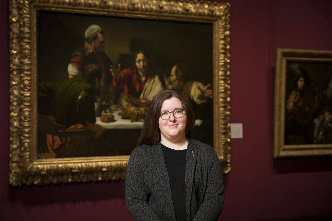 Siobhan Jolley is a white woman with long dark hair and round glasses, wearing a dark grey jacket over a black dress. She stands in the National Gallery in front of Caravaggio’s painting The Supper at Emmaus.