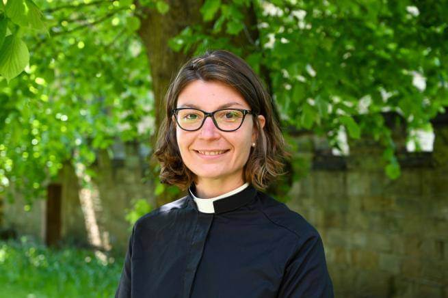 Stephanie is a white woman with shoulder length dark hair wearing dark rimmed glasses and a clerical collar under a black cassock, standing in front of a row of leafy green trees.