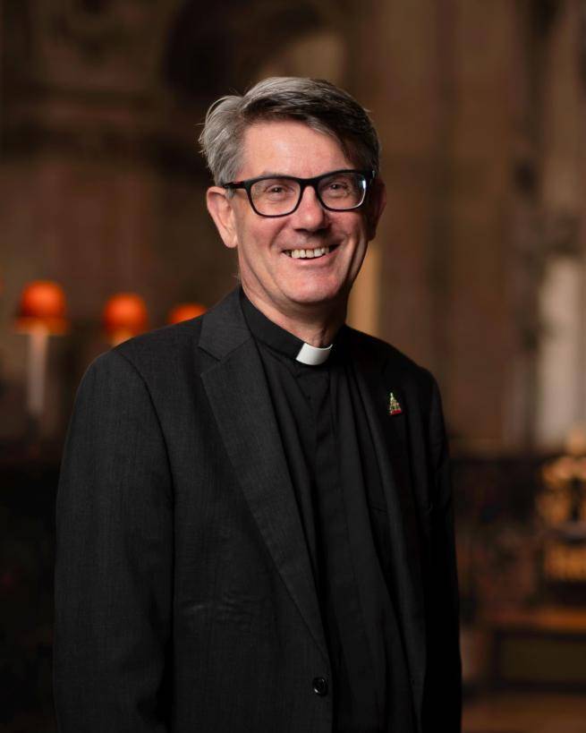 Andrew is a white man with short grey hair and black rimmed glasses wearing a clerical collar, black shirt and black jacket. The lamps of the quire of St Paul's in the background.