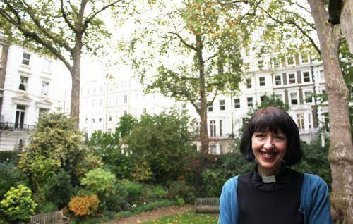 Carys, a white woman with short brown hair wearing a clerical collar, stands in a garden square in a city