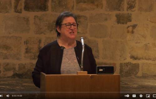 Gemma Simmonds, a white woman with glasses and short brown, speaking from the lectern in the Wren Suite, an exposed brick wall behind her.