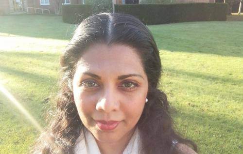 Sanjee Perera is a young Asian woman with long dark hair, photographed on a green lawn with sunlight across the lawn behind her, and a chapel in the background.