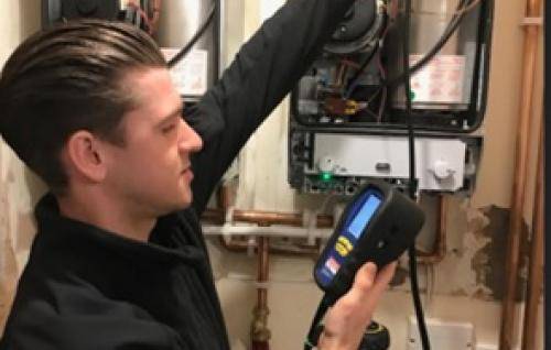 Oliver is a young white man with dark hair and wearing a black shirt. He is holding a piece of testing equipment which is attached to an open boiler.