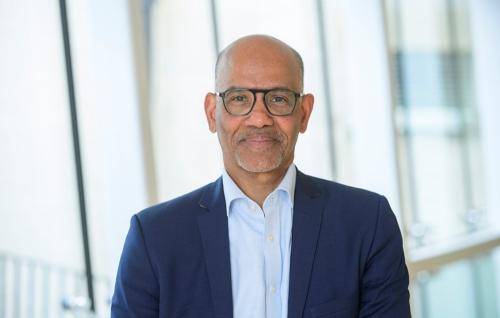 Alan Smith, a black man with bald head and black glasses wearing a navy blazer over a pale blue shirt, stands in front of large glass windows.