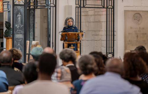 A black woman stands at a lectern speaking to a full audience