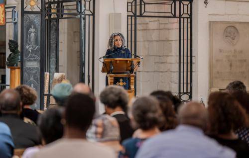 A black woman stands at a lectern speaking to a full audience