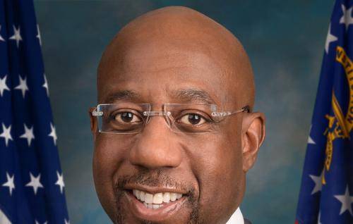 Raphael Warnock is a black man with a bald head, wearing frameless glasses, a dark suit with a white shirt and stripy tie. Behind him are two US flags.