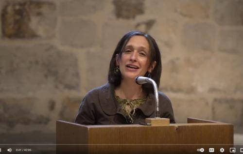 Susanna is a white woman with shoulder length dark hair standing at a microphone and lectern in front of an old stone wall inside the Cathedral