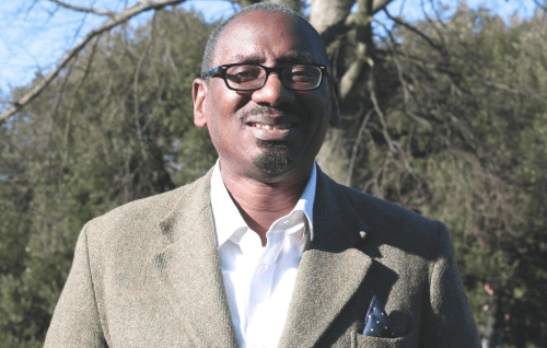 Richard is a black man with glasses and a bald head. He stands in a suit jacket and open necked white shirt in front of a wintery bare tree, with a pale blue sky