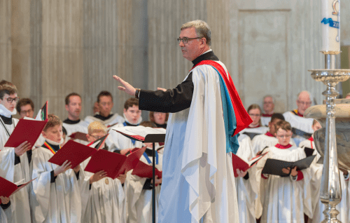 A white man with grey hair and glasses wearing a choir robe conducts a choir of choristers, also wearing white robes.