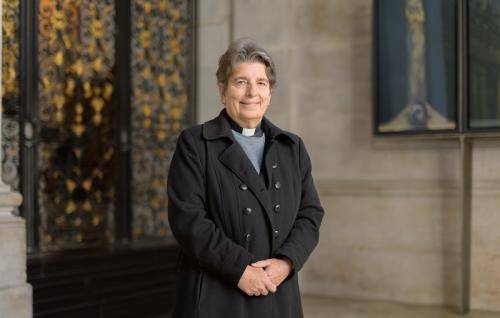 Paula Hollingsworth, a white woman with short dark hair in a clerical collar, stands in front of a wooden staircase.