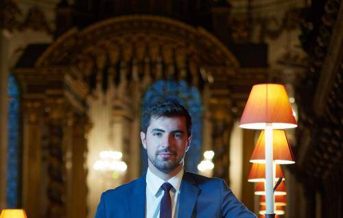 A portrait of James Orford. James is a white man with dark, short hair and a beard. He is wearing a suit and tie, and is stood in the Quire of St Paul's Cathedral.