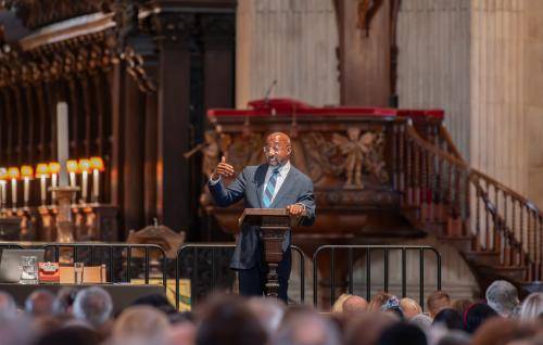 Raphael Warnock, a black man with bald head and glasses wearing a dark suit and blue striped tie, stands a t a lectern on stage in St Paul's Cathedral, the pulpit and quire in the background