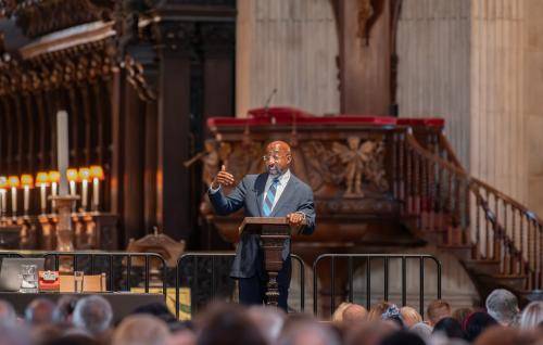 Raphael Warnock, a black man with bald head and glasses wearing a dark suit and blue striped tie, stands a t a lectern on stage in St Paul's Cathedral, the pulpit and quire in the background