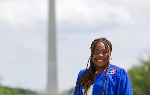 Alexandra is a young black woman wearing an academic robe standing in front of the Washington Monument and Lincoln Memorial Pool