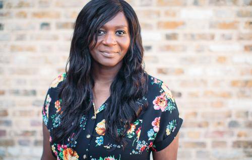 Chine is a black woman with long black hair wearing a floral patterned black dress and standing in front of a brick wall