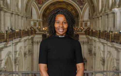 Catherine Okoronkwo is a black woman with shoulder length braided hair, wearing a short sleeved black top with a clerical collar. She stands in the Cathedral high up on the triforium level, with the nave of the Cathedral behind her.