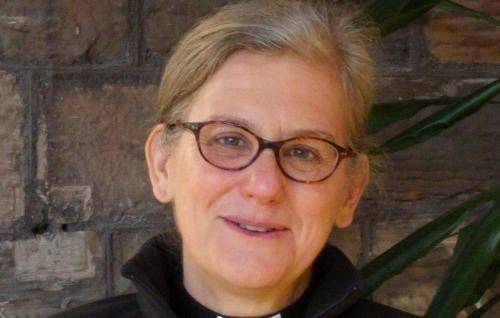 Janet Williams is a white woman with short grey hair and dark rimmed glasses wearing a clerical collar under a black jacket