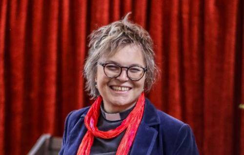 Rachel is a white woman with short grey hair and glasses wearing a clerical collar under a blur top and jacket with a red scarf. She stands alongside a high-sided wooden chair and in front of a deep red velvet curtain