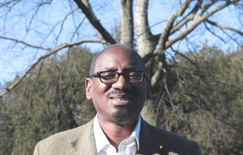 Richard is a black man with a bald head and dark rimmed glasses wearing a beige suit jacket over a white shirt, standing outdoors in front of a bare tree and a pale blue sky