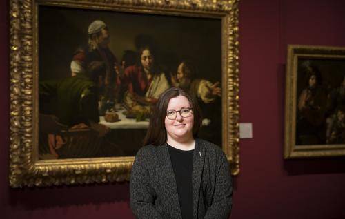 Siobhan Jolley is a white woman with long dark hair and round glasses, wearing a dark grey jacket over a black dress. She stands in the National Gallery in front of Caravaggio’s painting The Supper at Emmaus.