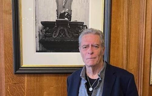 A white man with short grey hair stands in a wood panelled room by a large photo of St Paul's pulpit with a man preaching