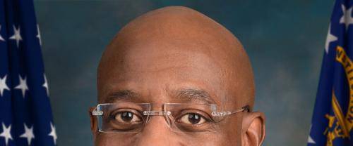 Raphael Warnock is a black man with a bald head, wearing frameless glasses, a dark suit with a white shirt and stripy tie. Behind him are two US flags.