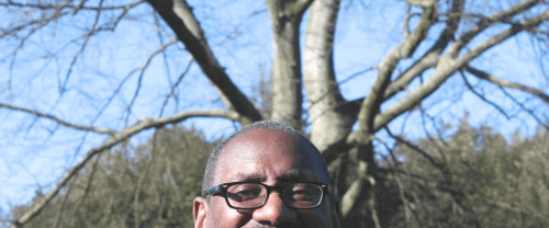 Richard is a black man with glasses and a bald head. He stands in a suit jacket and open necked white shirt in front of a wintery bare tree, with a pale blue sky