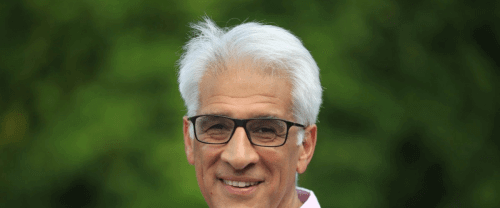 Steve is a mixed race man with white hair and black glasses, wearing a pink shirt, and standing outside with out of focus green foliage behind him.